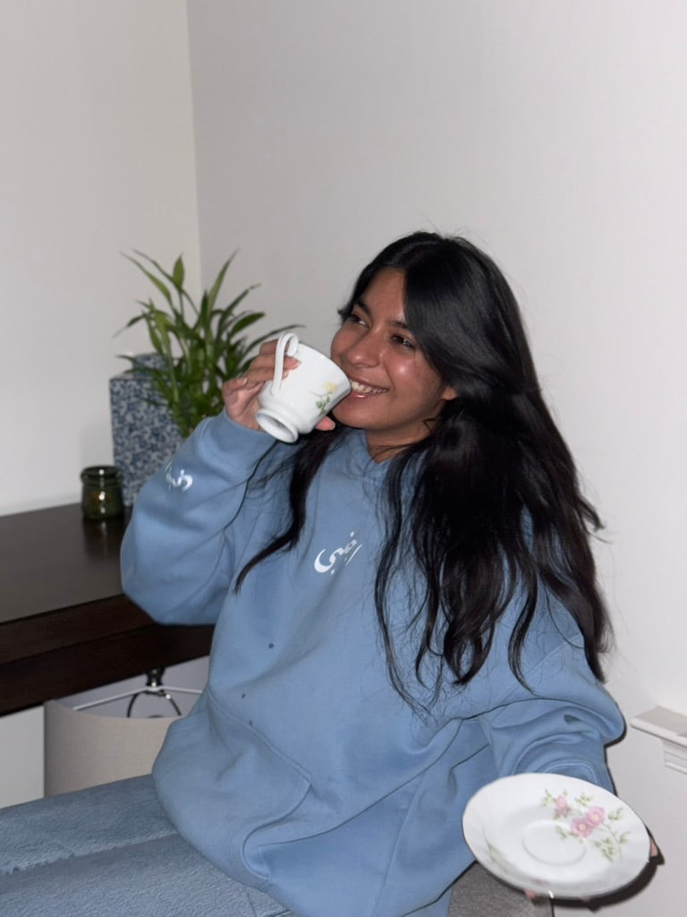 Woman in a blue sweatshirt drinking from a mug in a casual setting.