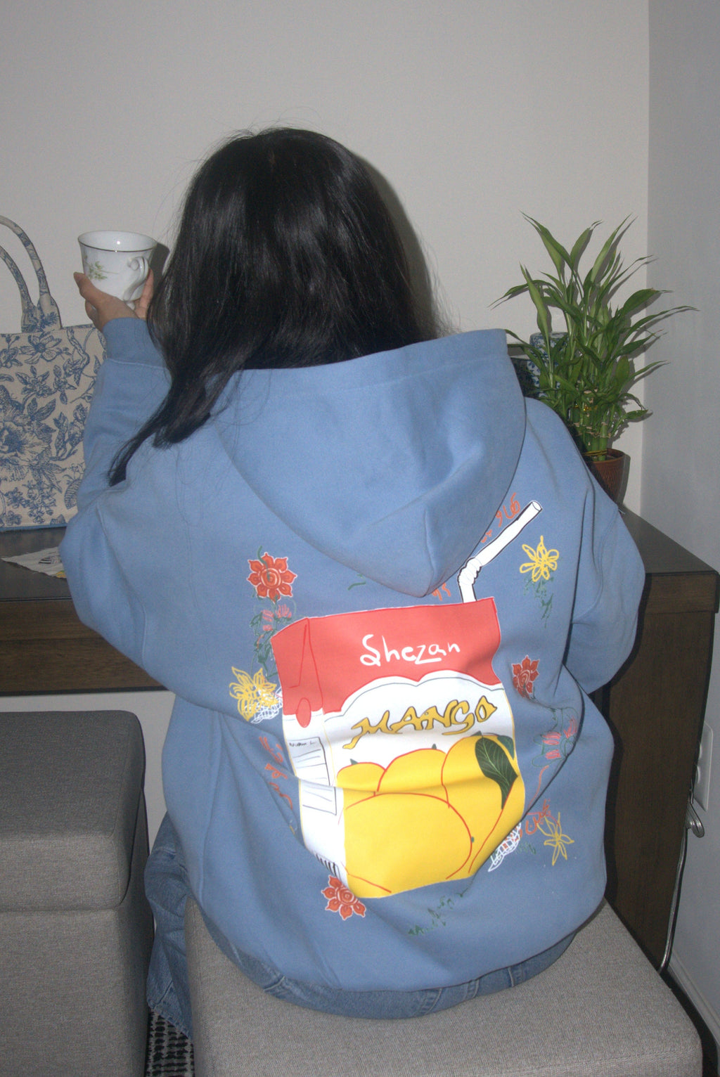 Person wearing a blue pakistani mango juice hoodie with a colorful design sitting on a gray ottoman in a room.